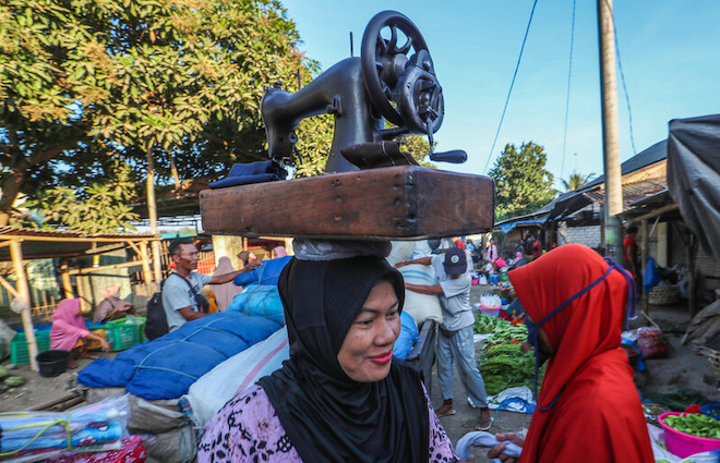 A woman with carrying a hand-cranked sewing machine walks through a traditional market in Indonesia.
