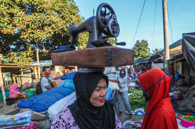 A woman with a portable sewing machine walks through a traditional market in Indonesia.