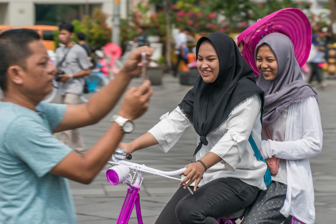 A man taking a photo using his mobile phone while two men ride a bike in a popular tourist destination in Indonesia.