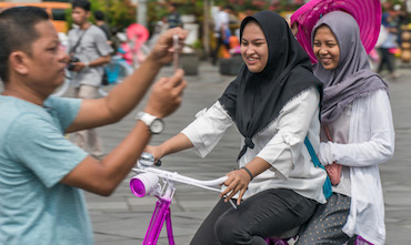 A man taking a photo using his mobile phone while two men ride a bike in a popular tourist destination in Indonesia.