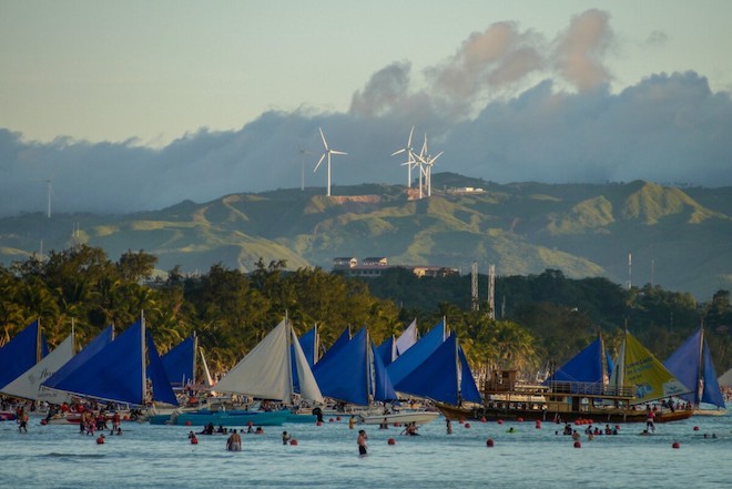 Wind turbines are seen in the background of a crowded beach.