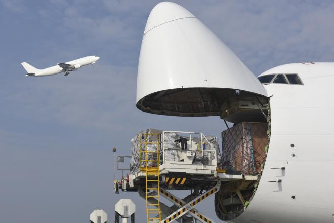 A plane flies over a jet with the nose open for cargo loading.