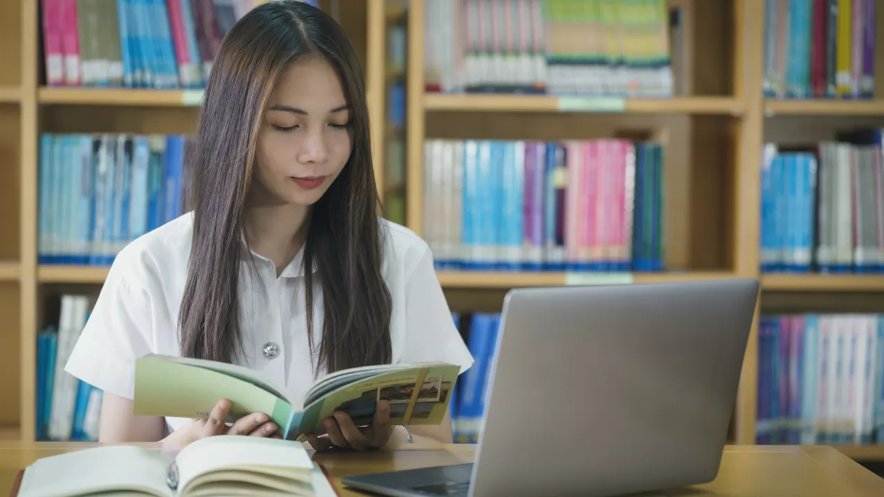 A woman reading a book with a laptop in front of her on the  table. 